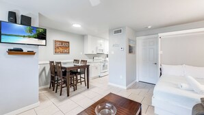 Una sala moderna con un televisor de pantalla plana, un comedor con mesa y sillas de madera, una cocina con armarios blancos y un dormitorio con cama blanca y una puerta.