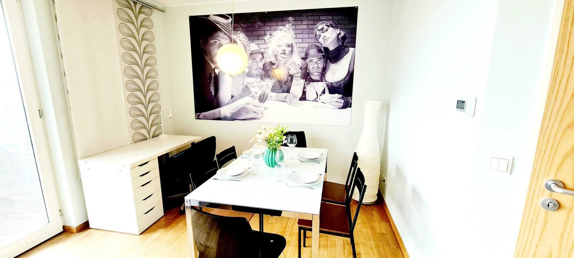 Ein Essbereich mit einem weißen Tisch, gedeckt für vier Personen, einer Blumenvase und einem großen Poster an der Wand.