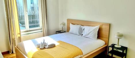 Una cama bien hecha con tablero de madera, una mesita de noche con lámpara y una ventana con cortinas.
