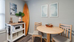 A dining area with a wooden table and chairs, a white microwave on a white cabinet, a surfboard leaning against the wall, and two framed artworks. - Cozy Studio Large Pool w/ a Private Courtyard and a 5 Min Walk to the Beach (Port Aransas)