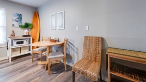 A dining area with a wooden table and chairs, a surfboard, and a woven chair. - Cozy Studio Large Pool w/ a Private Courtyard and a 5 Min Walk to the Beach (Port Aransas)