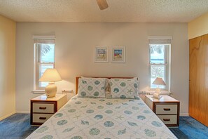 Un dormitorio con una cama, dos mesitas de noche, un ventilador de techo, dos ventanas con persianas y dos cuadros en la pared.
