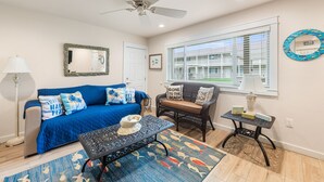 A living room with a blue sofa, a coffee table, a wicker chair, a small table with a lamp, and a large window with blinds.