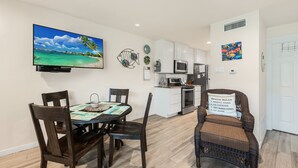 A dining area with a table and chairs, a kitchen in the background, and a beach-themed pillow on a chair.