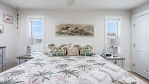 A bedroom with a bed, two nightstands, a ceiling fan, a painting, and two windows with blinds.