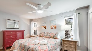 Un dormitorio con cama, cómoda roja, mesita de noche, ventilador de techo y ventana con cortinas.