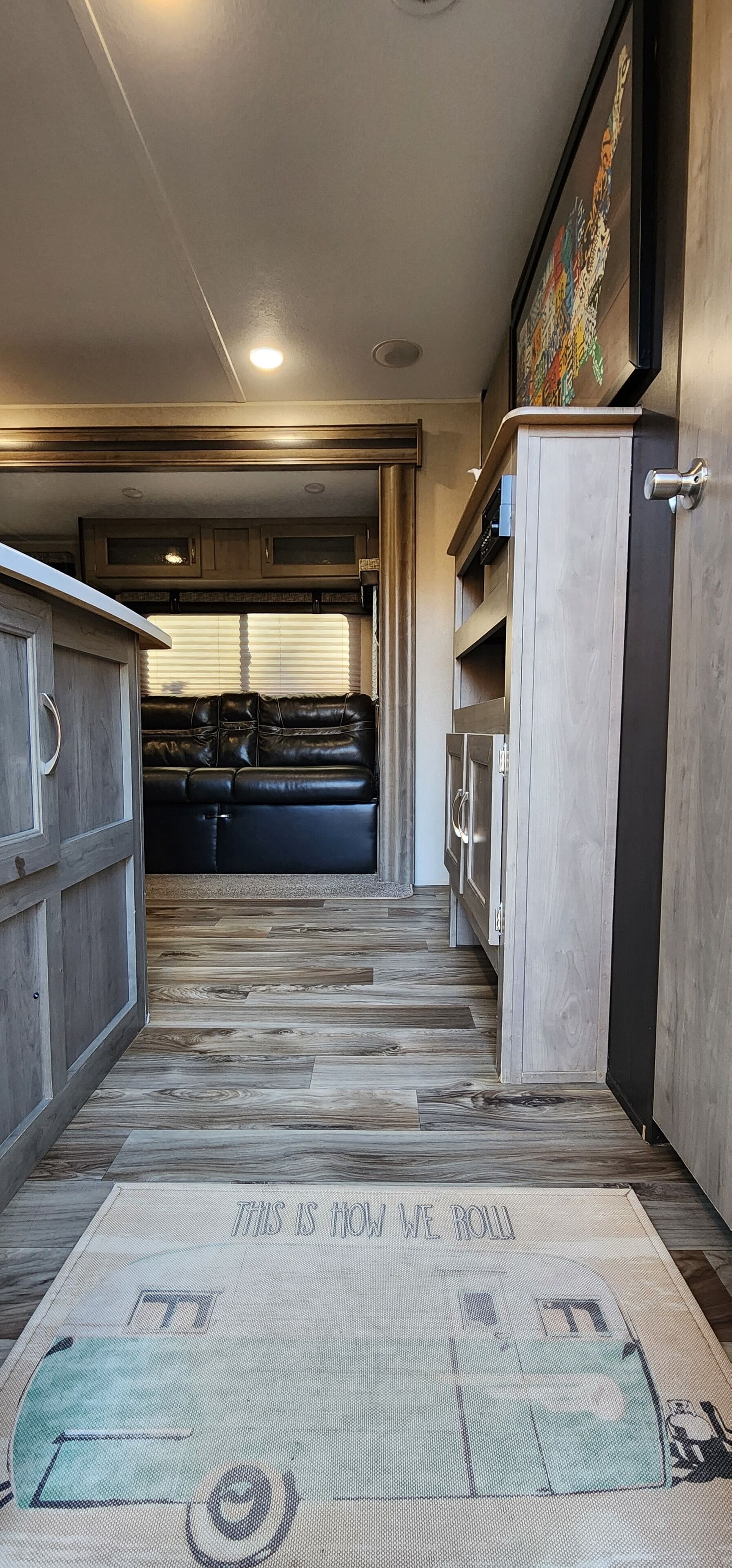 A narrow hallway with a wooden floor leading to a seating area with a black leather couch. There are cabinets and a door on the right side.