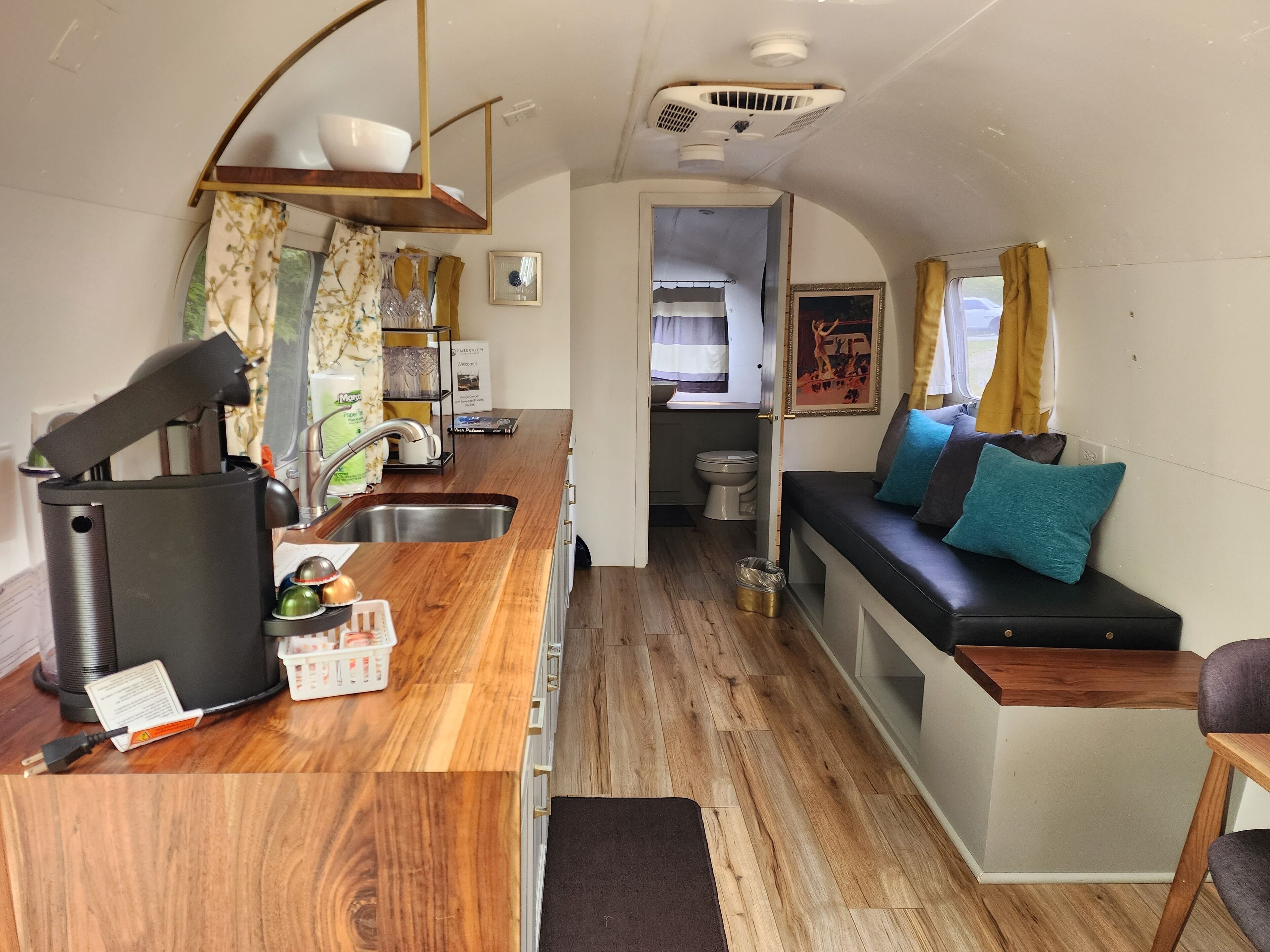 A compact kitchen with a sink, a coffee maker, and a small countertop. There is a bench with cushions and a window with curtains.
