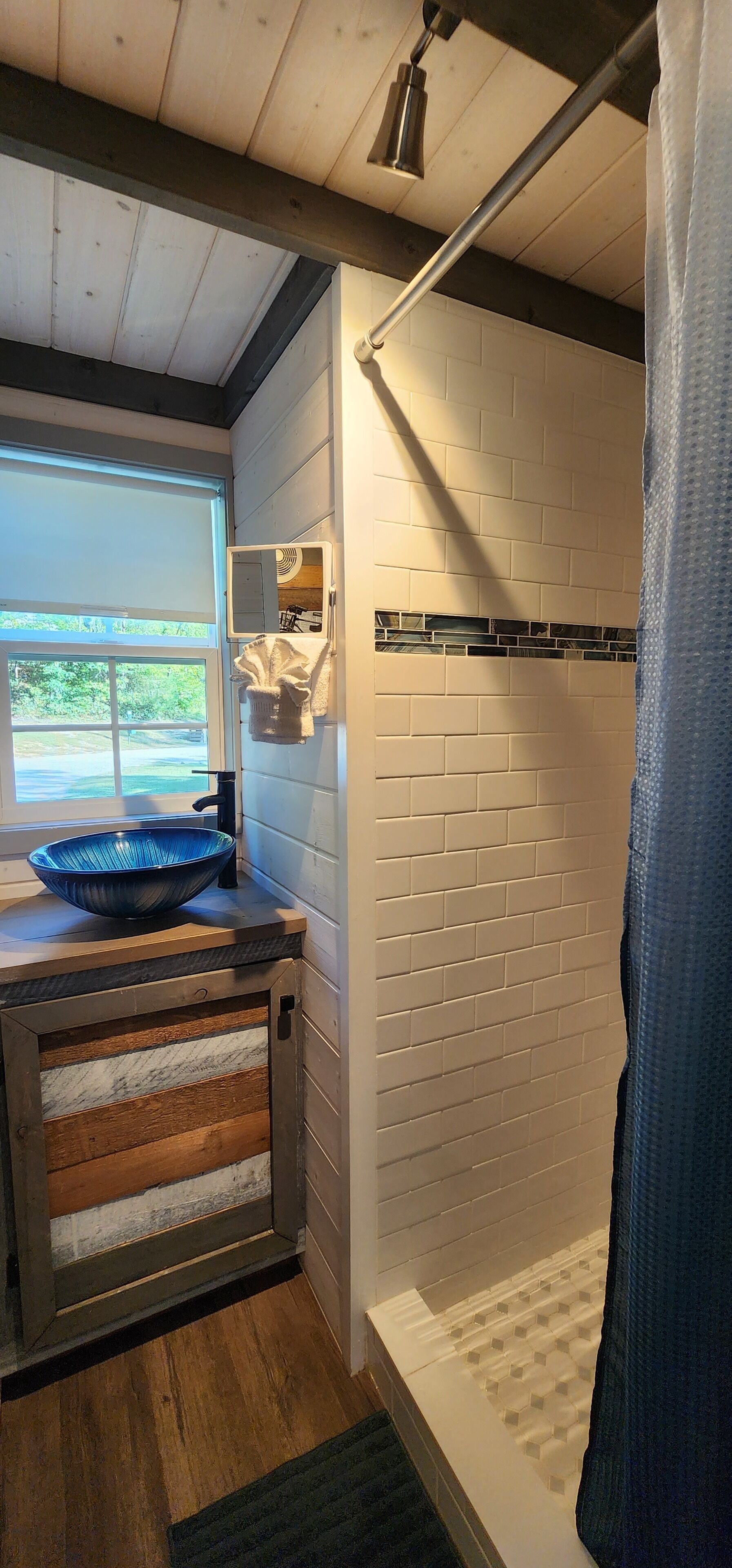 A bathroom with a wooden floor, a blue basin, a white tiled shower area, and a window with a view of greenery.