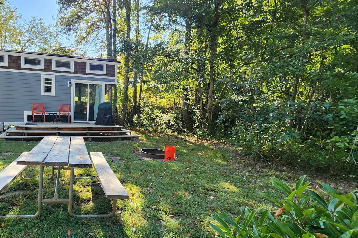 A small cabin with a deck, two red chairs, and a picnic table in a forested area.