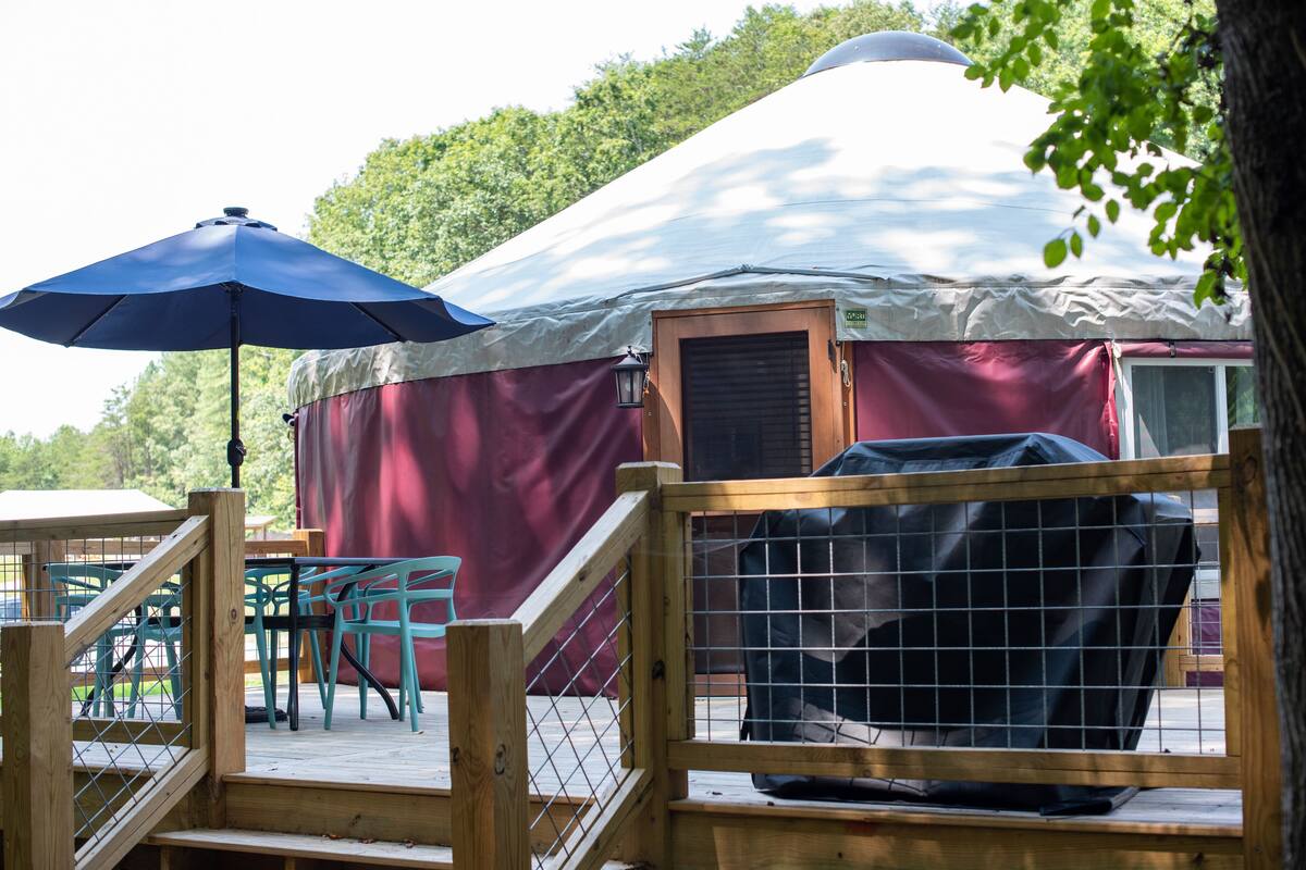 A yurt with a wooden entrance, a covered grill, and a blue umbrella.