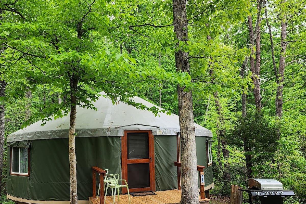 A green A-frame yurt with a wooden deck, a door, and a grill.