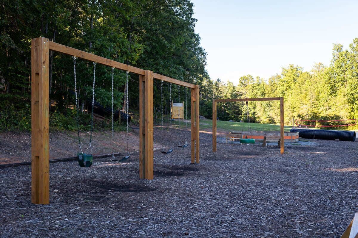 A wooden swing set in a park with a gravel ground and trees in the background.