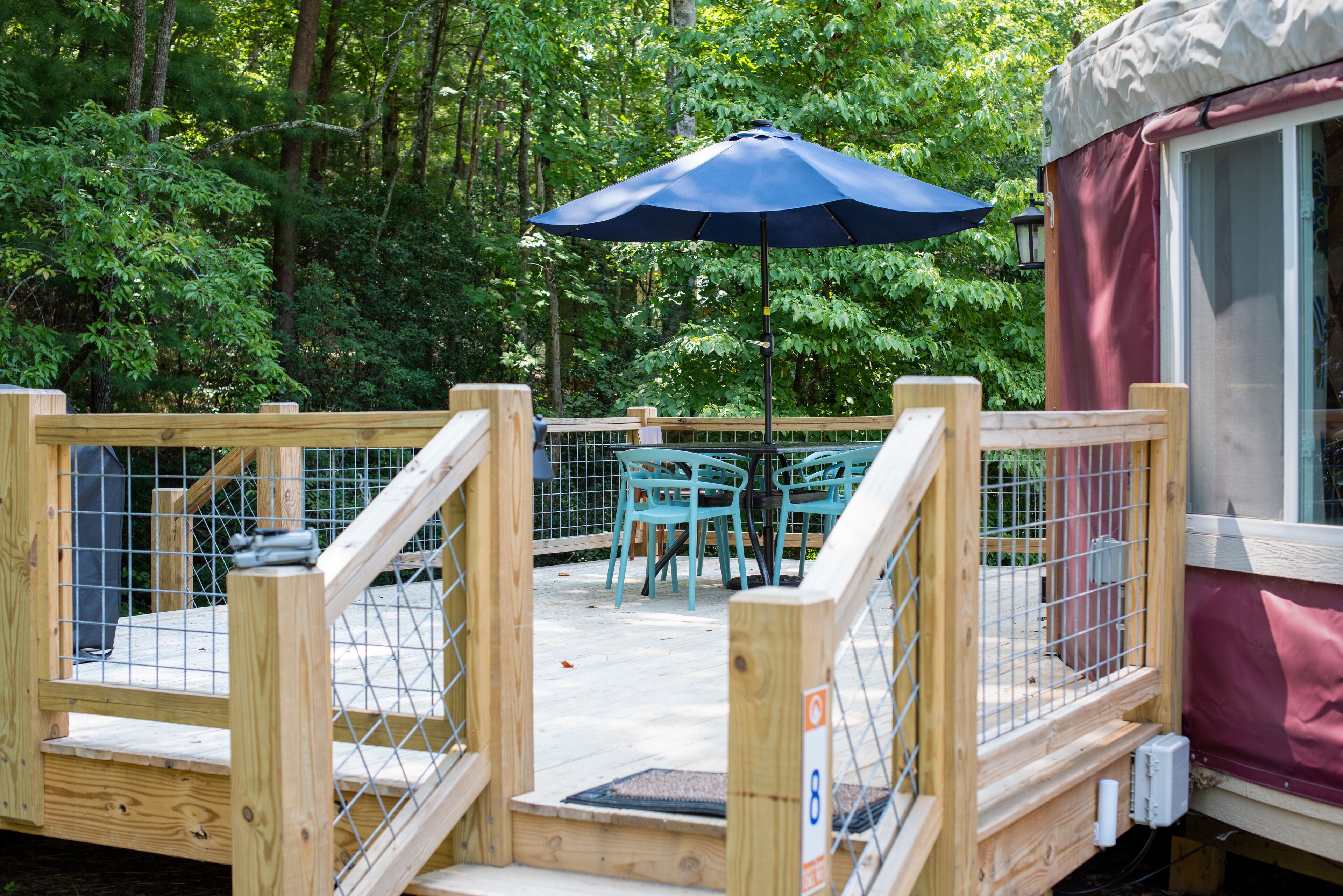 A wooden deck with a blue umbrella, turquoise chairs, and a wooden fence.