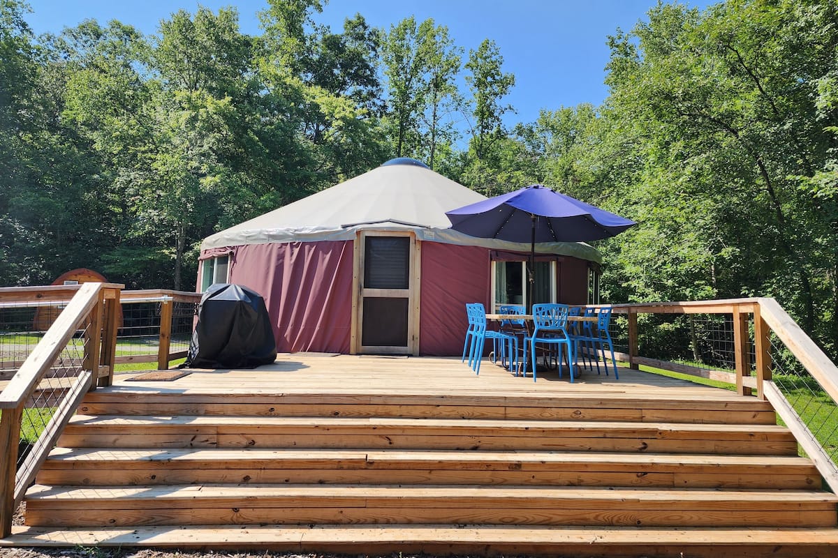 A yurt with a covered deck, blue chairs, and a barbecue grill.