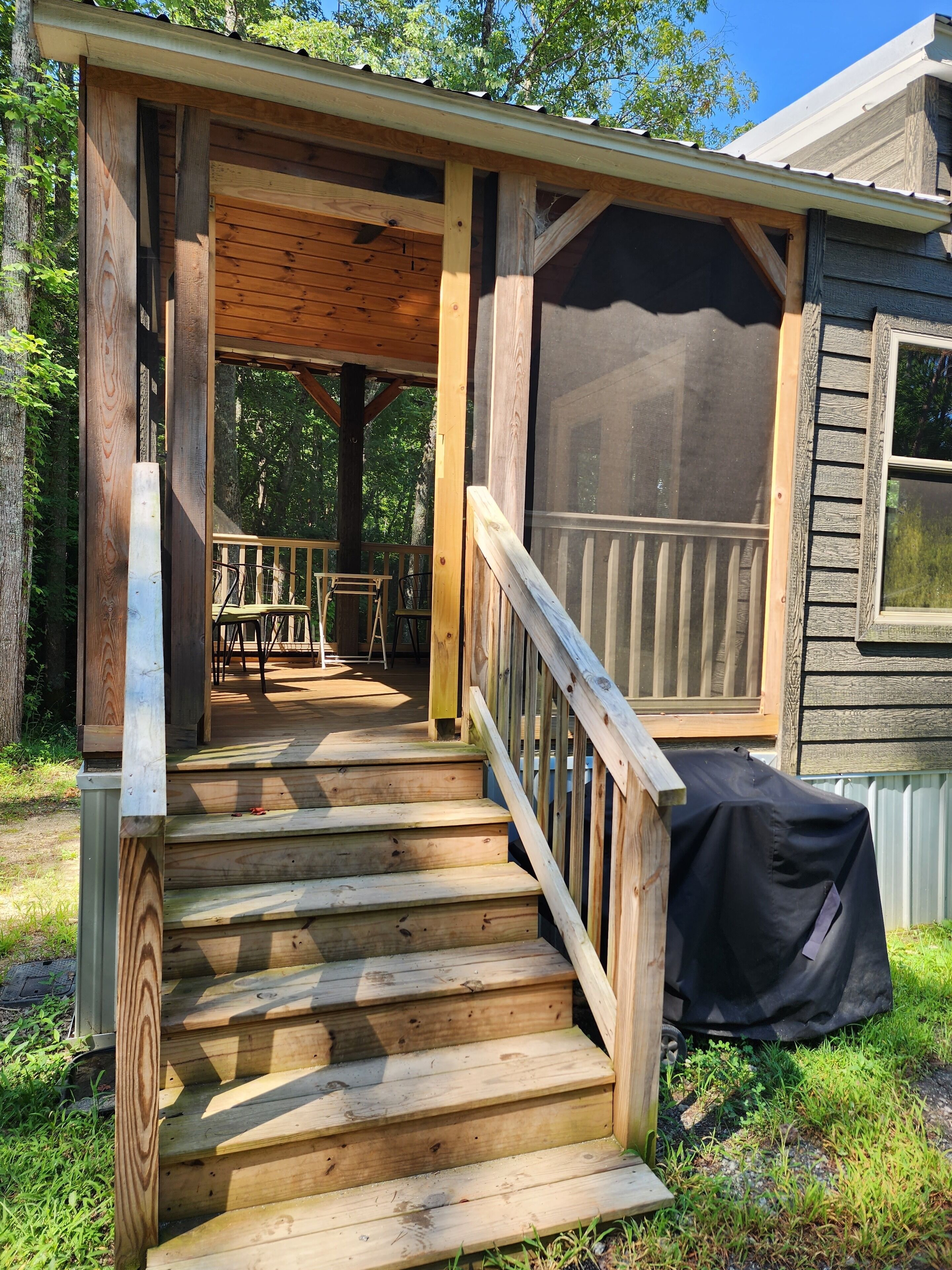 A wooden staircase leading to a covered porch with a railing, surrounded by greenery.