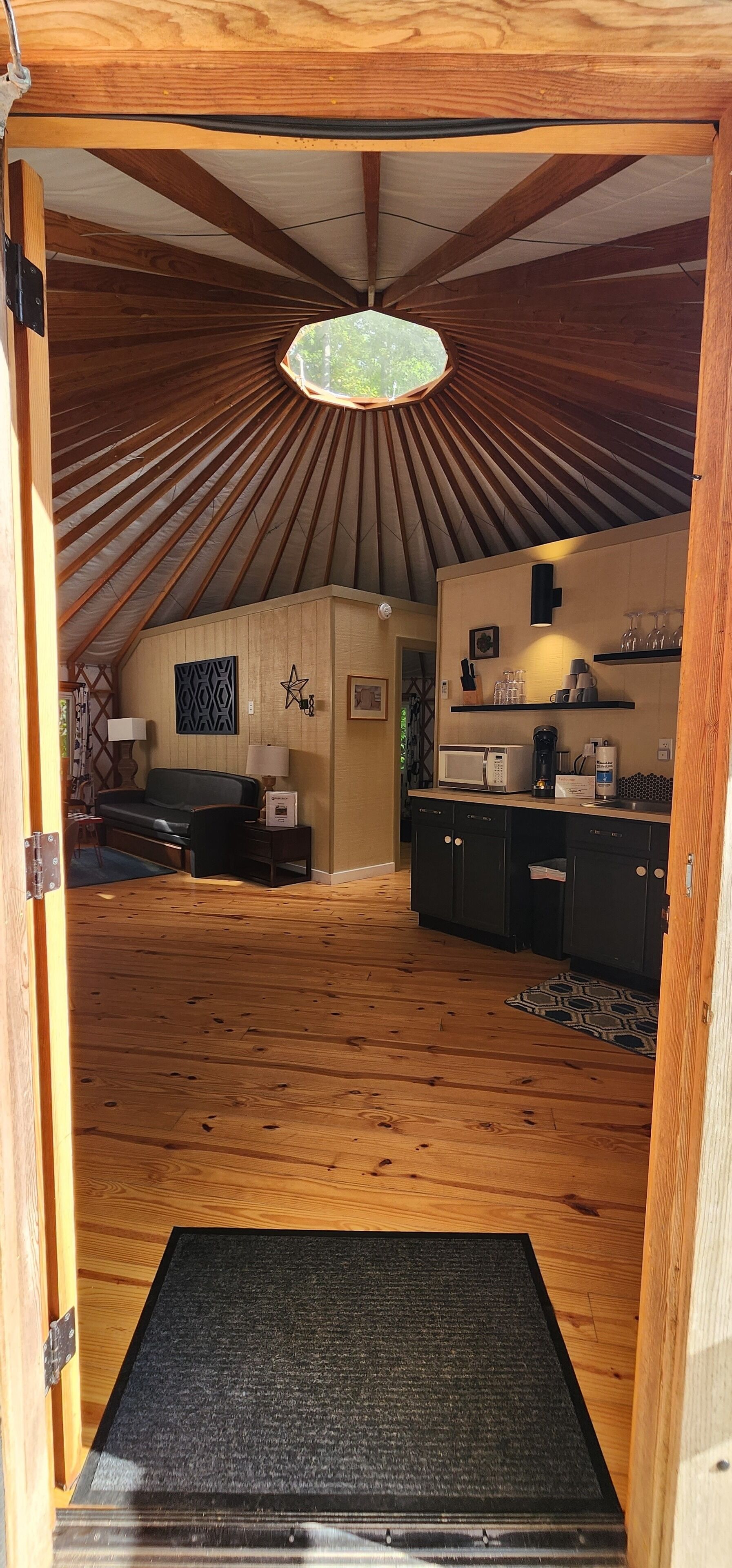 An interior with a wooden floor, a kitchen area with cabinets and appliances, a dining area with a table and chairs, and a ceiling with wooden beams and a hanging light fixture.