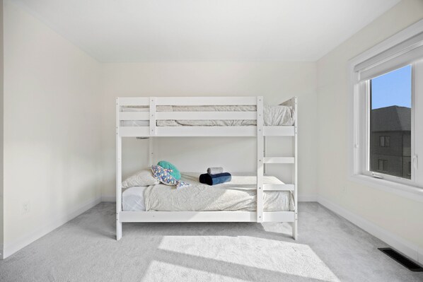 Uma cama de solteiro em um quarto com janela, piso coberto de carpete e um respiradouro visível.