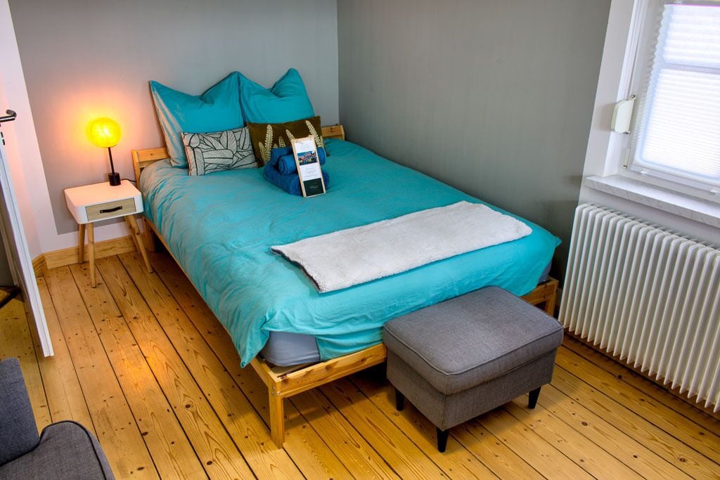 Een slaapkamer met een bed, nachtkastje, lamp, bankje en een raam.