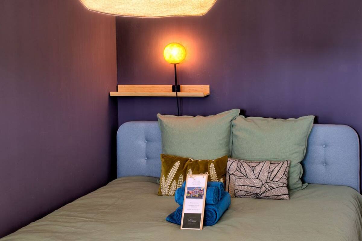 Una cama bien hecha con un cabecero azul, dos almohadas y una toalla doblada, con una lámpara sobre ella.