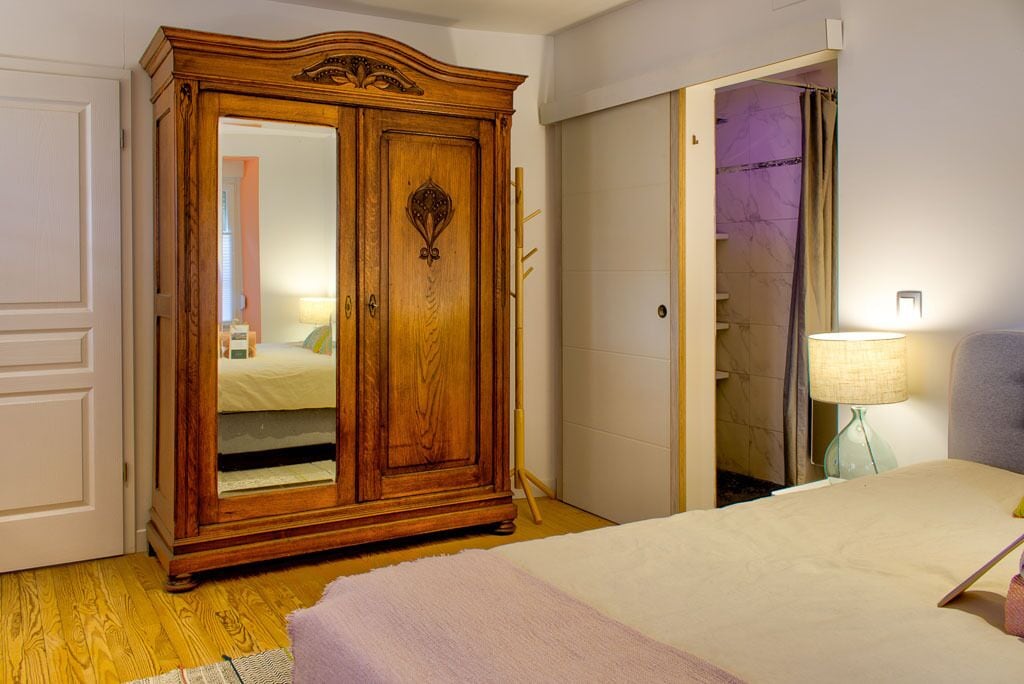 A bedroom with a wooden wardrobe, a bed with a white duvet, a bedside lamp, and a bathroom visible through an open door.