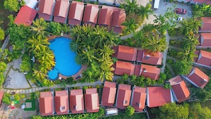Vista aérea de un complejo turístico con una piscina central rodeada de palmeras y edificios con techos rojos.
