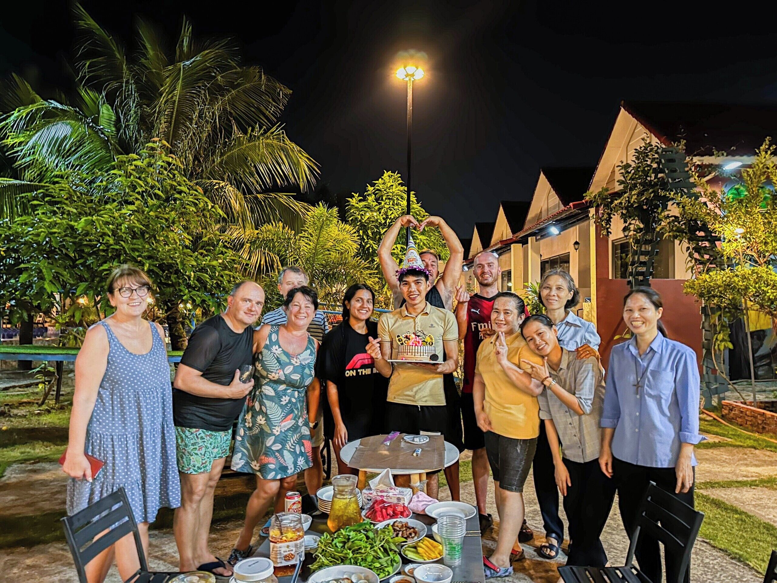 A group of people gathered around a table with food and drinks, outdoors at night.