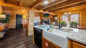 A wooden cabin interior with a kitchen featuring a granite countertop, a dishwasher, and a stone fireplace. - Sevierville Cabin, Hot Tub & Mountain Views (Sevierville)