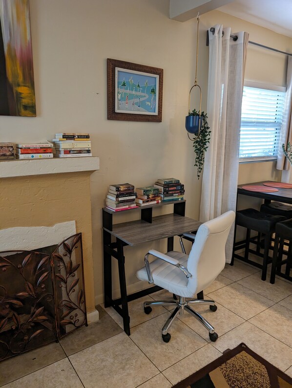 Un despacho en casa con una silla blanca, un escritorio con libros, un cuadro enmarcado y una ventana con cortinas.