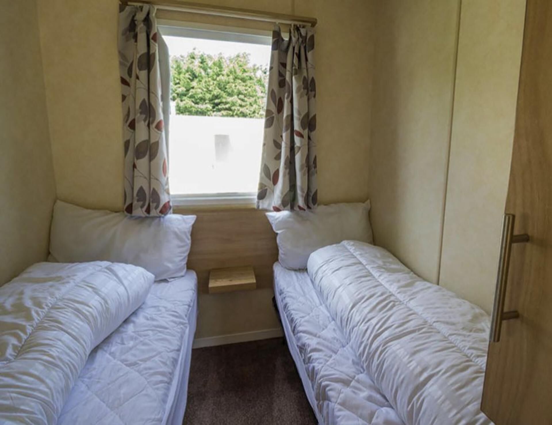 Ein kleiner Wohnmobil mit einem Schlafzimmer, zwei Betten, einem Fenster mit Vorhängen und einer hölzernen Bank in der Mitte.