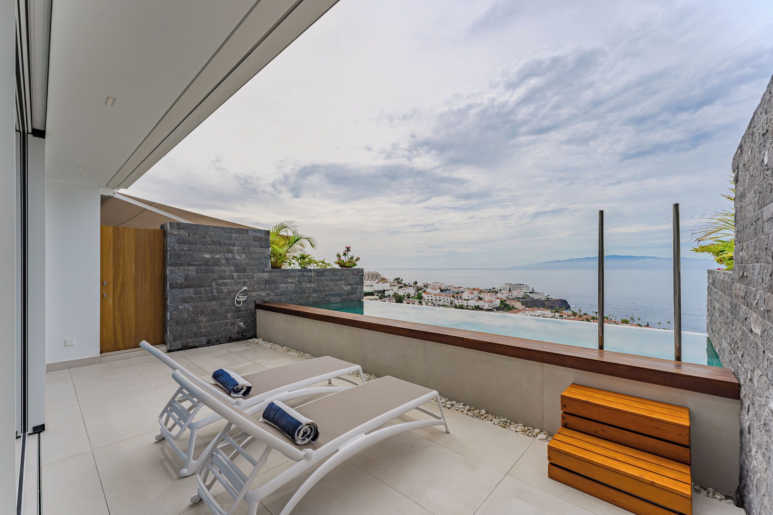 A modern balcony with a view of the sea, a pool, and a wooden bench.