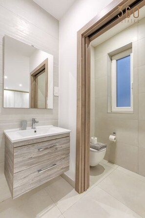 A modern bathroom with a wooden vanity, a large mirror, and a toilet with a window above it. - Modern 3BR home with Stunning Marina View in Pieta (Tal-Pietà)