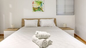 A neatly made bed with white linens and rolled towels, two white pillows, wooden headboard, bedside tables with lamps, and a colorful abstract painting on the wall. - Lovely Studio home in Prime area of Sliema w/WIFI (Sliema)