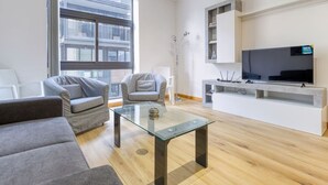 Ein modernes Wohnzimmer mit Glastisch, grauen Sofas, einem Flachbildfernseher und einem großen Fenster mit Blick auf die Stadt.