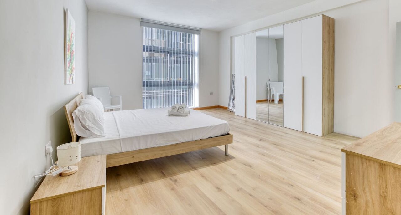 Ein schlicht eingerichtetes Schlafzimmer mit einem hölzernen Bett, einem Nachttisch und einem großen Fenster mit Jalousien.