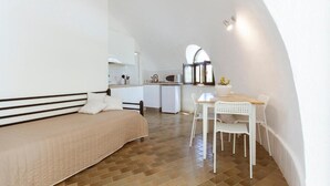 A bedroom with a bed, a small table, and a chair. There is a kitchenette in the background.