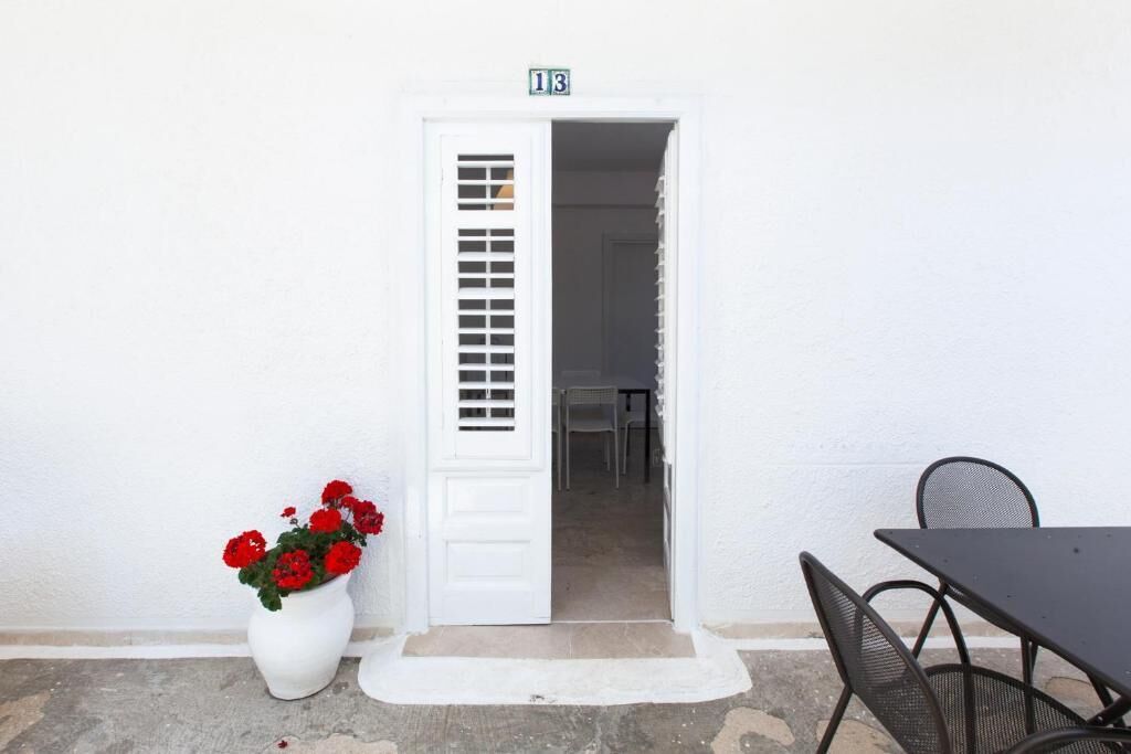Une porte blanche avec une petite fenêtre, un pot de fleurs avec des fleurs rouges et une table avec des chaises devant un bâtiment.