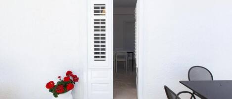 Une porte blanche avec une petite fenêtre, un pot de fleurs avec des fleurs rouges et une table avec des chaises devant un bâtiment.