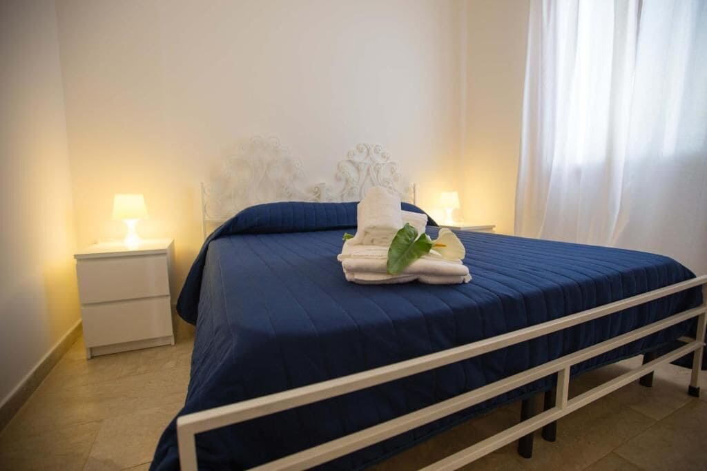 Cama bem arrumada com colcha azul e cabeceira branca, duas luminárias de cabeceira e uma mesa de cabeceira branca.