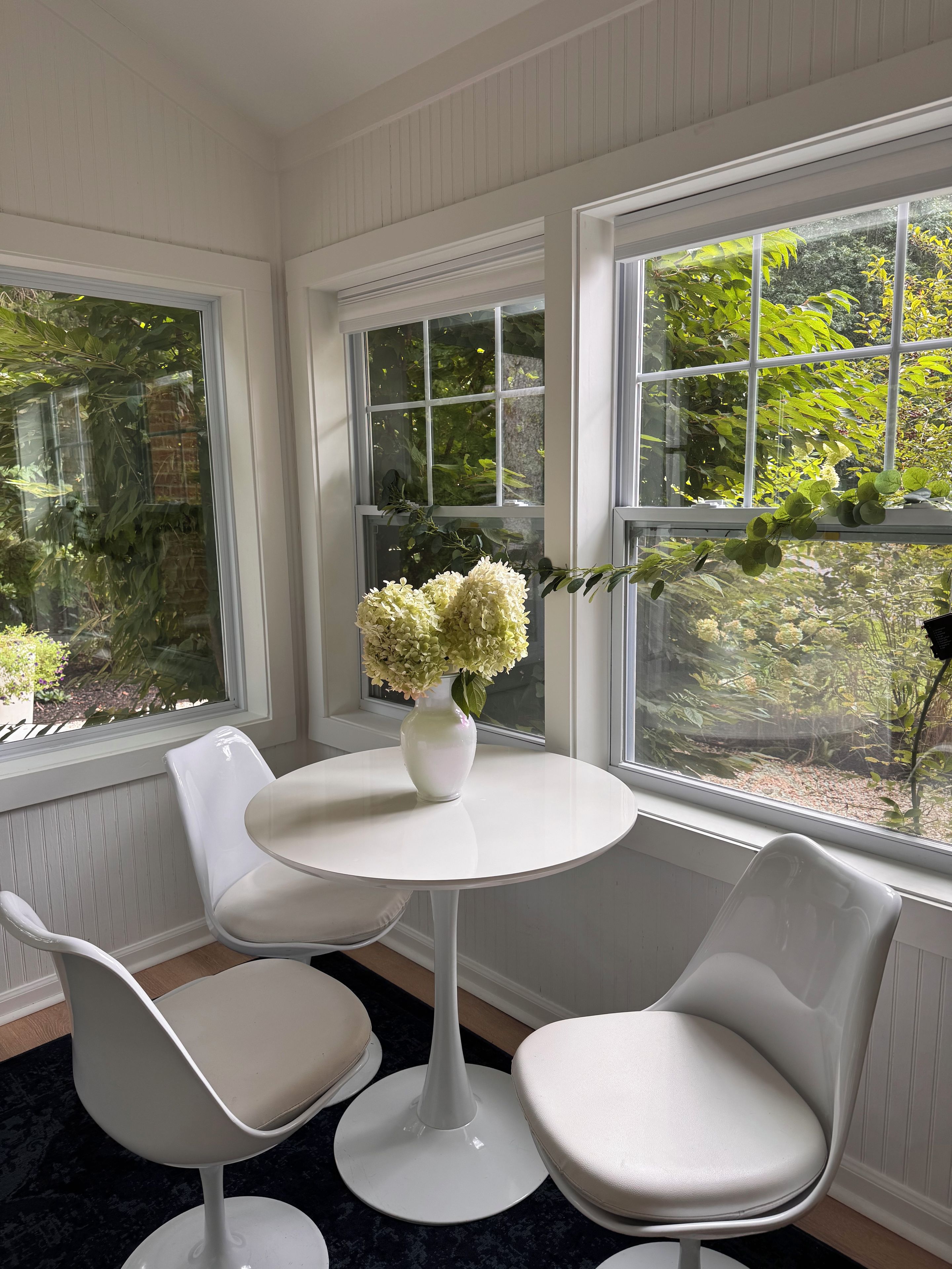 Une pièce avec une table ronde blanche, deux chaises blanches et un vase rempli de fleurs.