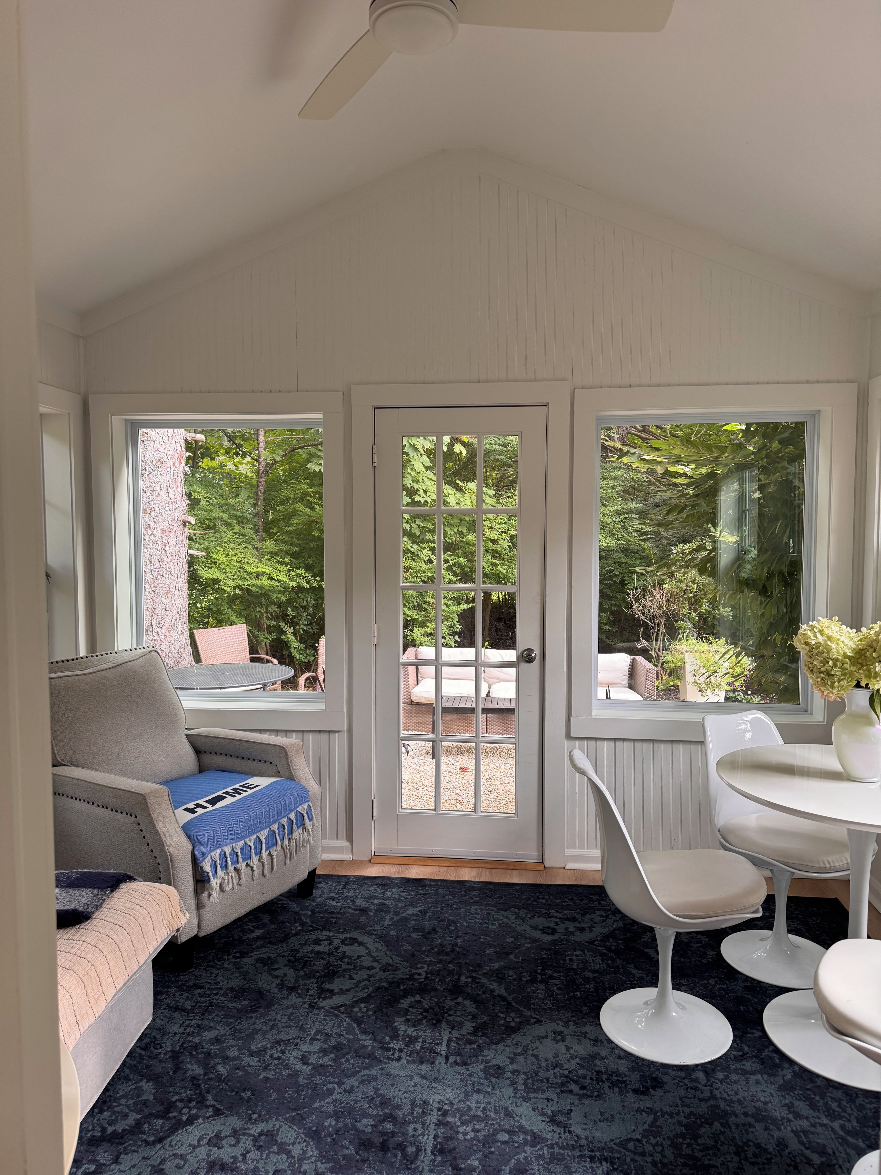 Une pièce avec un ventilateur de plafond, une porte donnant sur l’extérieur, un canapé avec un coussin bleu, une table ronde avec un vase et un tapis à motifs.