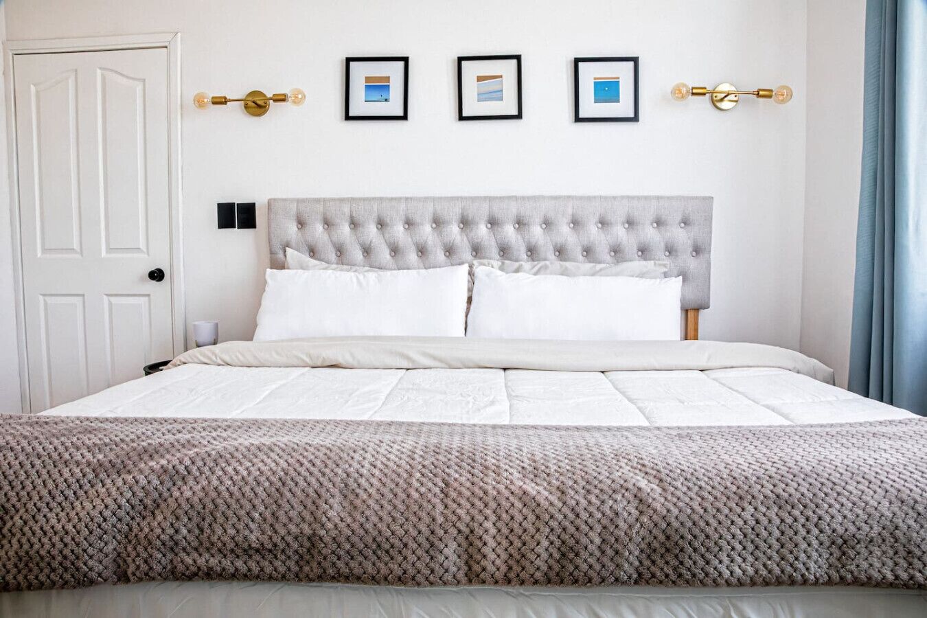 A neatly made bed with a grey tufted headboard, white bedding, and a patterned throw blanket. There are three framed pictures on the wall above the bed.