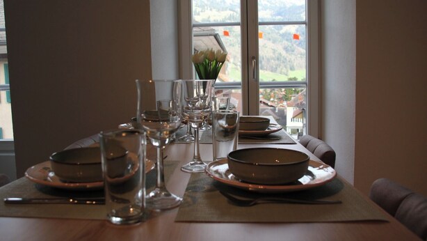 Une table à manger dressée avec des assiettes, des bols et des verres, offrant une vue sur les montagnes par la fenêtre.