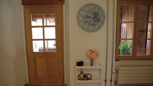 Une porte en bois avec une fenêtre, une horloge murale, une petite étagère blanche ornée d’un vase de fleurs et un radiateur.
