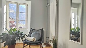 Une pièce avec un plancher en bois, une chaise noire avec un coussin, une plante en pot, un radiateur et une fenêtre donnant sur les montagnes.