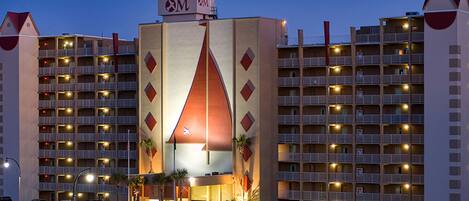 A hotel with a distinctive triangular central tower, multiple balconies, and a parking lot with various cars.