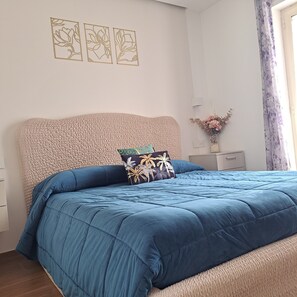 Un dormitorio con cama, cabecera, mesita de noche, un florero con flores y una ventana con cortinas.