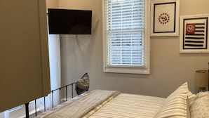 A bedroom with a bed, a window with blinds, a flat-screen TV mounted on the wall, and framed artwork on the wall. - Fantastic Location (Ogunquit)