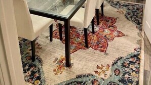 Comedor con mesa de vidrio, sillas blancas, alfombra estampada y vista a través de una puerta de cristal.