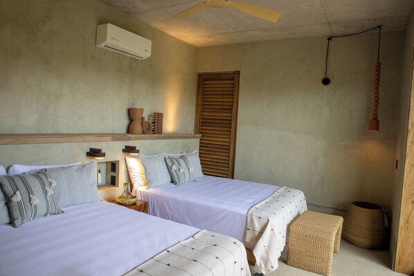 Een kamer met twee bedden, een plafondventilator, een aan de muur bevestigde airconditioner, een bedlampje en een geweven mand.
